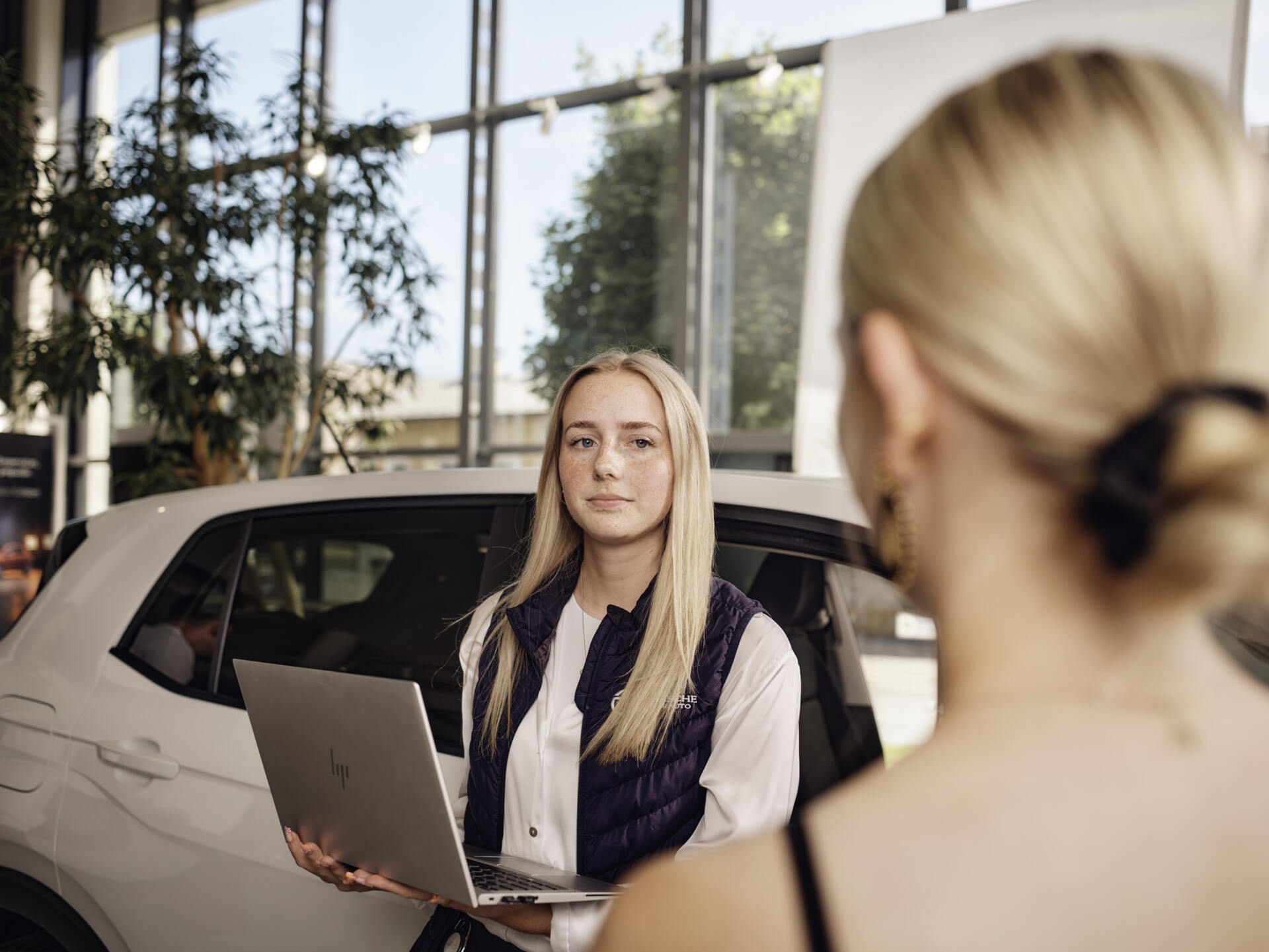 Verkäuferin mit Laptop in Autohaus vor weißem Auto und Fensterfront