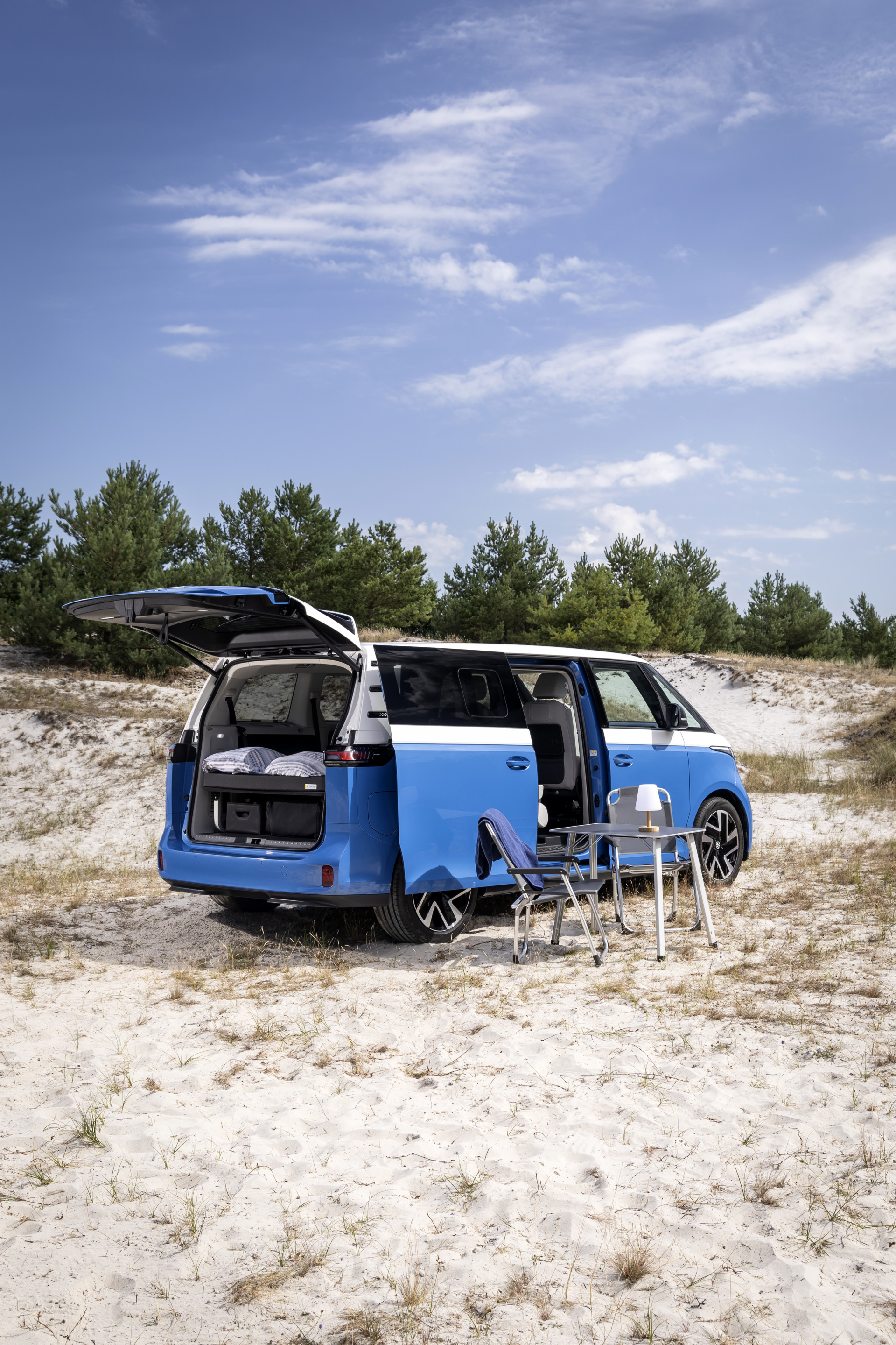 Blauer Camper mit geöffneter Heckklappe und Türen steht im Sand, daneben ein Tisch mit Stühlen.