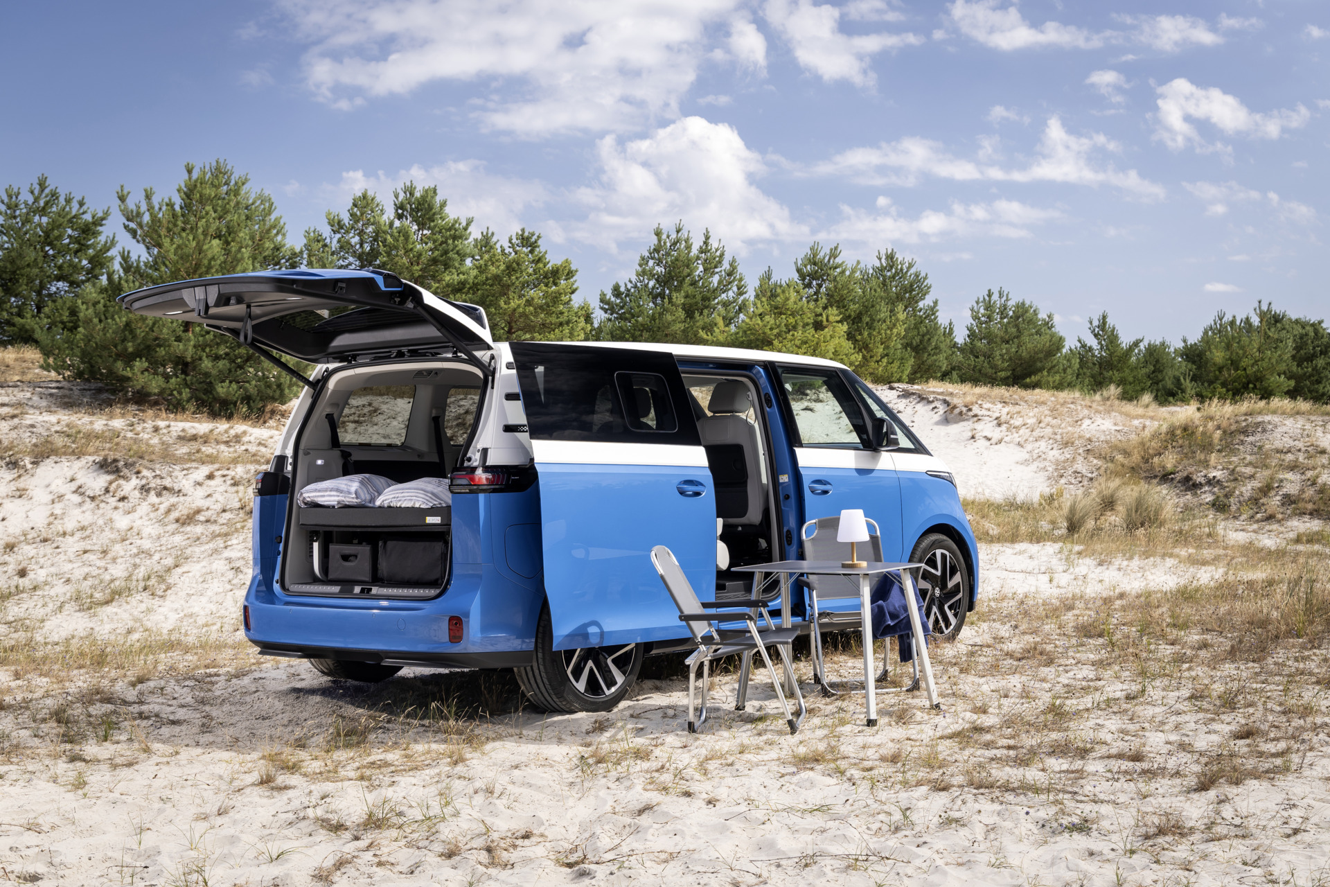 Blauer Campervan mit geöffneter Heckklappe und seitlicher Tür, Tisch und Stühlen im Sand, Pinienwald im Hintergrund