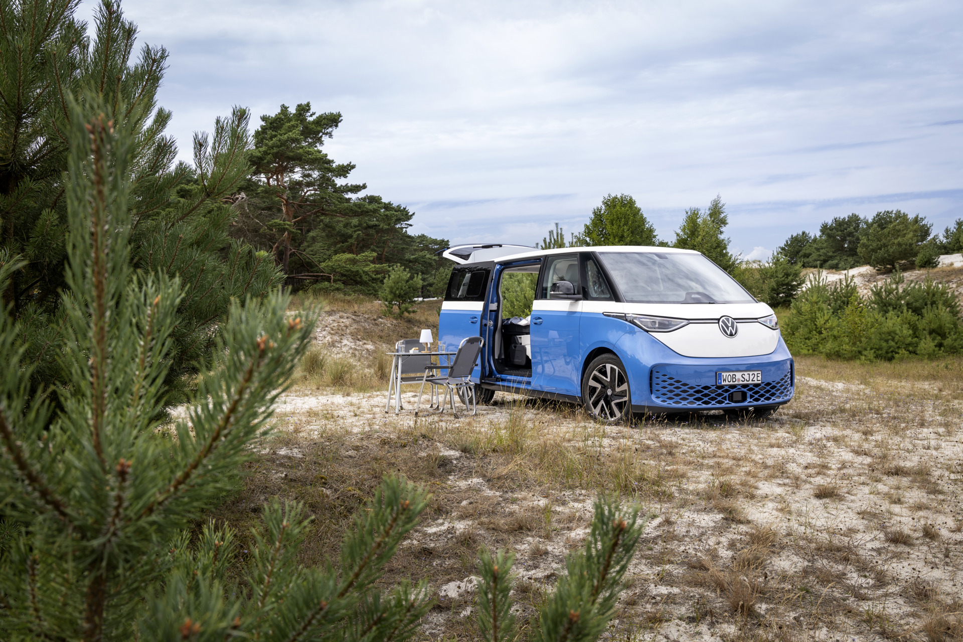 VW ID. Buzz Camper mit geöffneten Türen und Tisch mit Stühlen in einer sandigen Landschaft mit Kiefern.